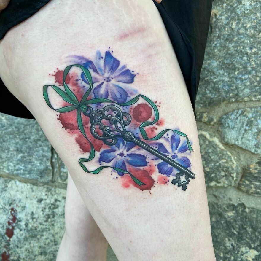 Watercolor tattoo of a vintage key with colorful flowers on a person's leg.