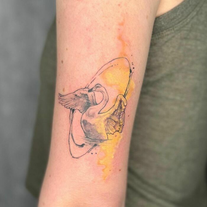 Watercolor tattoo of a swan with wings spread, surrounded by a splash of yellow and orange ink on a person's arm.