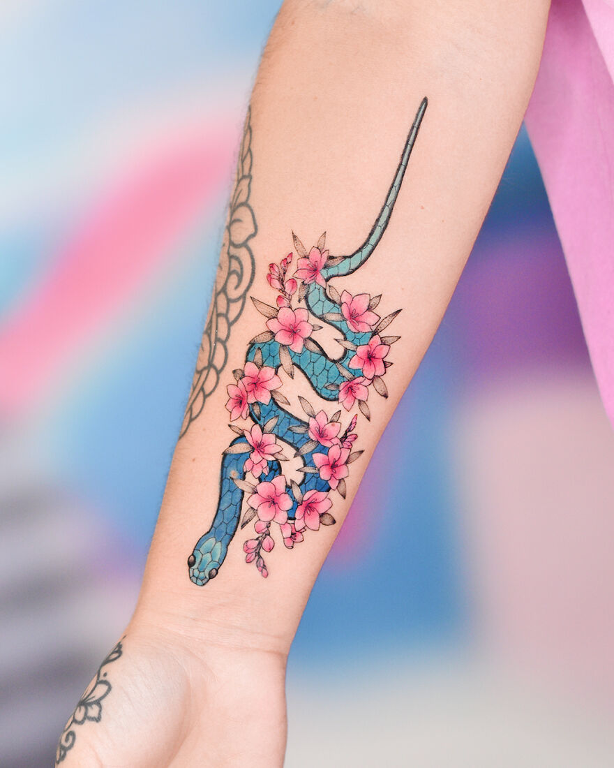 Watercolor tattoo of a blue snake intertwined with pink flowers on forearm.