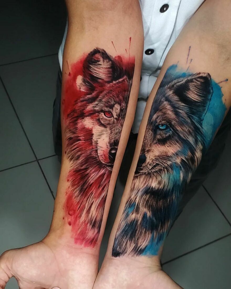 Two forearms with watercolor tattoos of wolf faces in red and blue.
