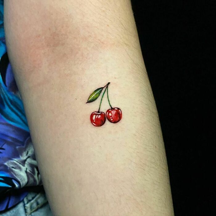 Watercolor tattoo of cherries with a leaf on an arm, showcasing vibrant colors and ethereal design.