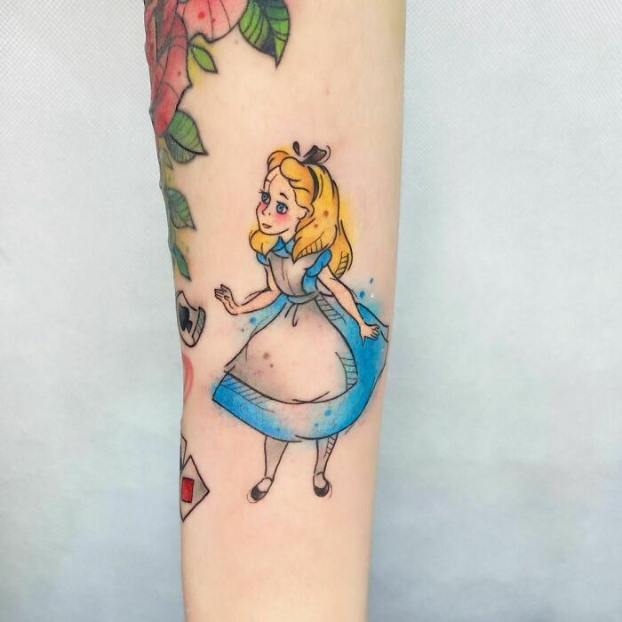 Watercolor tattoo of a whimsical character in a blue dress with floral accents on a forearm.