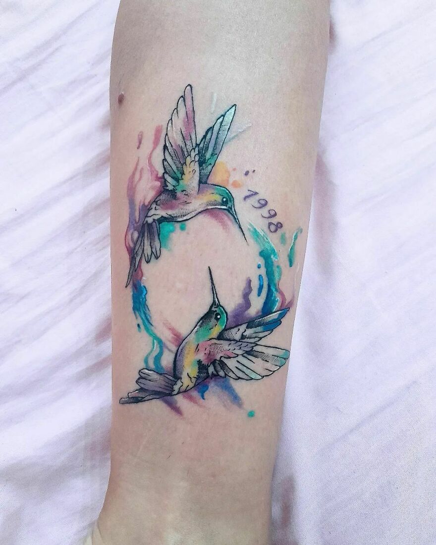 Watercolor tattoo of two hummingbirds in vibrant colors on forearm.