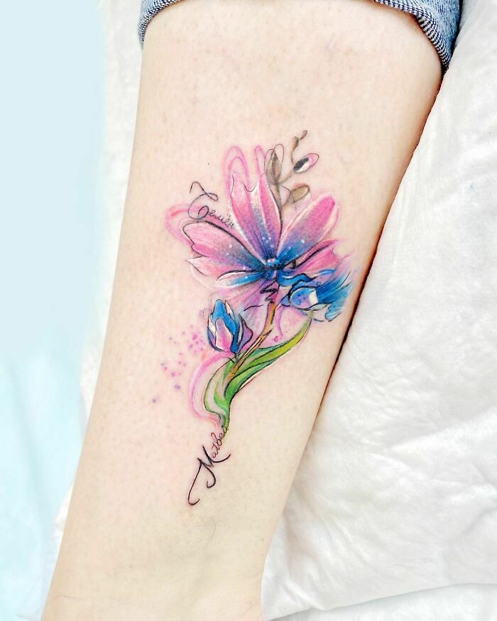 Ethereal watercolor tattoo of pink and blue flowers on forearm, blending vibrant hues and delicate designs.