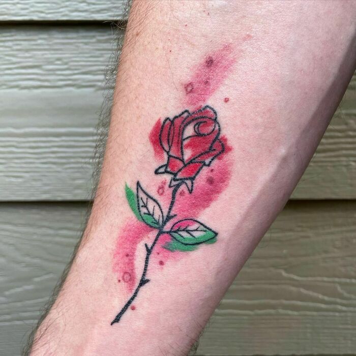 Watercolor tattoo of a rose on an arm, featuring pink and green splashes for inkspiration.