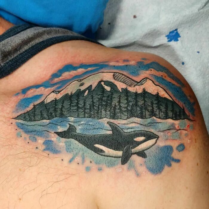 Watercolor tattoo of an orca swimming beneath a mountain landscape.