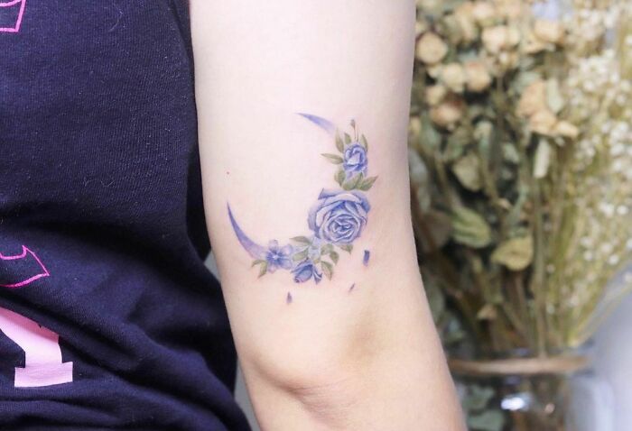 Watercolor tattoo of a crescent moon with blue roses and green leaves on an arm.