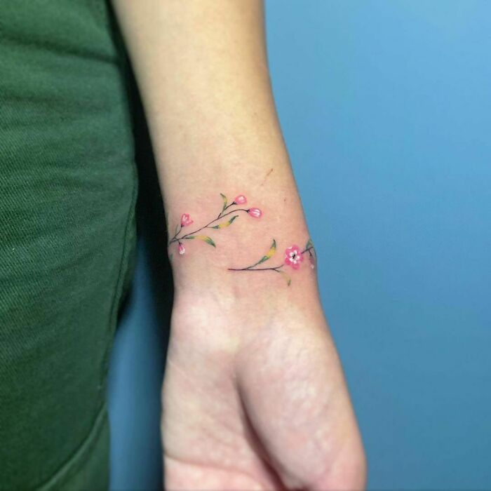 Delicate floral watercolor tattoo on wrist, showcasing pink blossoms and green leaves, offering inkspiration.