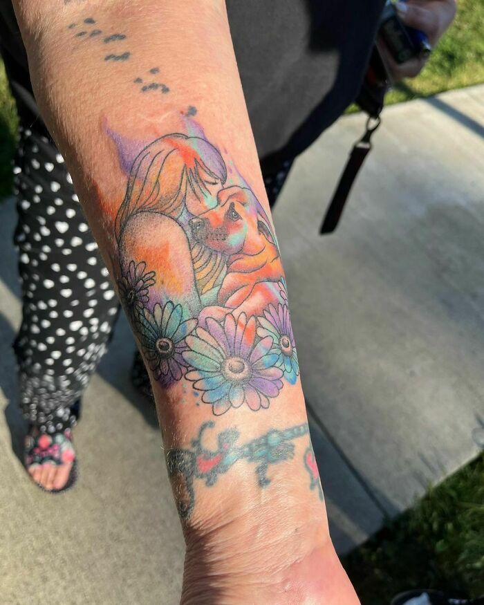 Watercolor tattoo of a woman and dog surrounded by colorful flowers on a forearm.