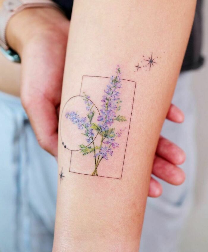Watercolor tattoo of purple flowers with geometric lines and stars on forearm.