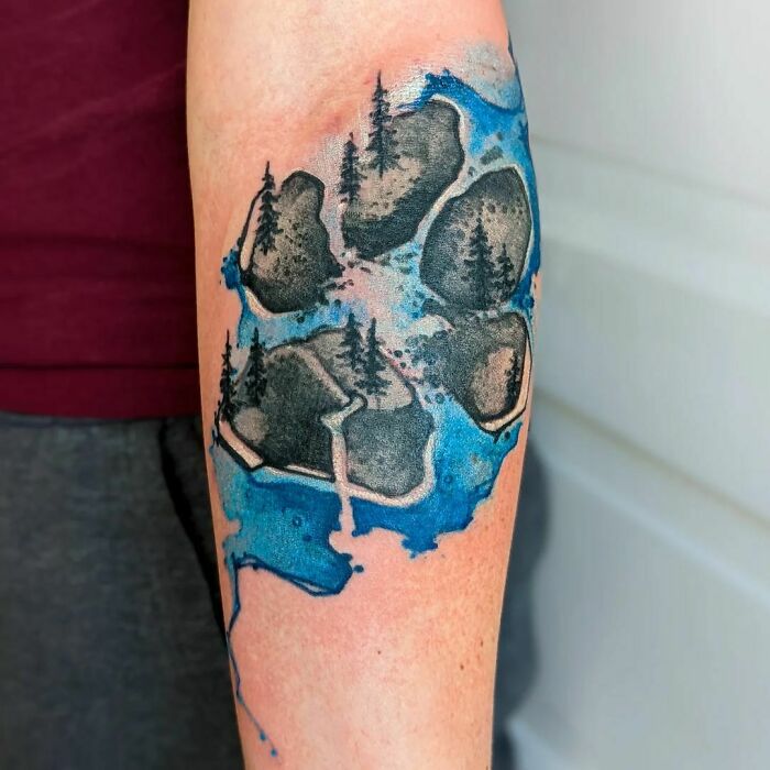 Watercolor tattoo with trees and rocks on forearm, featuring blue and black tones for an ethereal look.