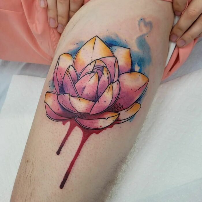 Watercolor tattoo of a vibrant lotus with splashes of blue and red ink on skin.