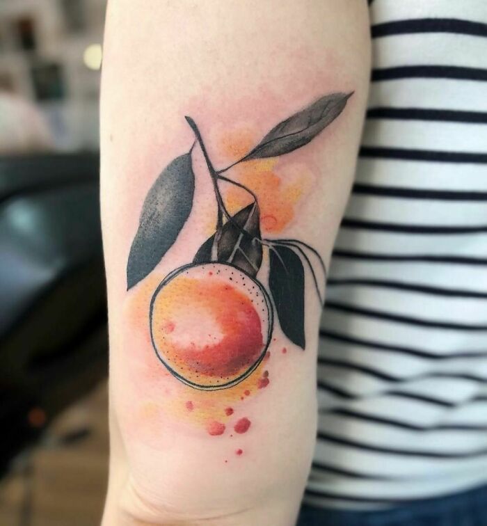 Watercolor tattoo of a vibrant orange with leaves, inked on an arm, showcasing ethereal artistry.