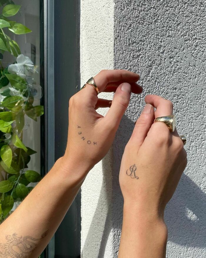 Two hands with word tattoos "Honey" and letter "R," adorned with rings, against a textured wall and greenery.