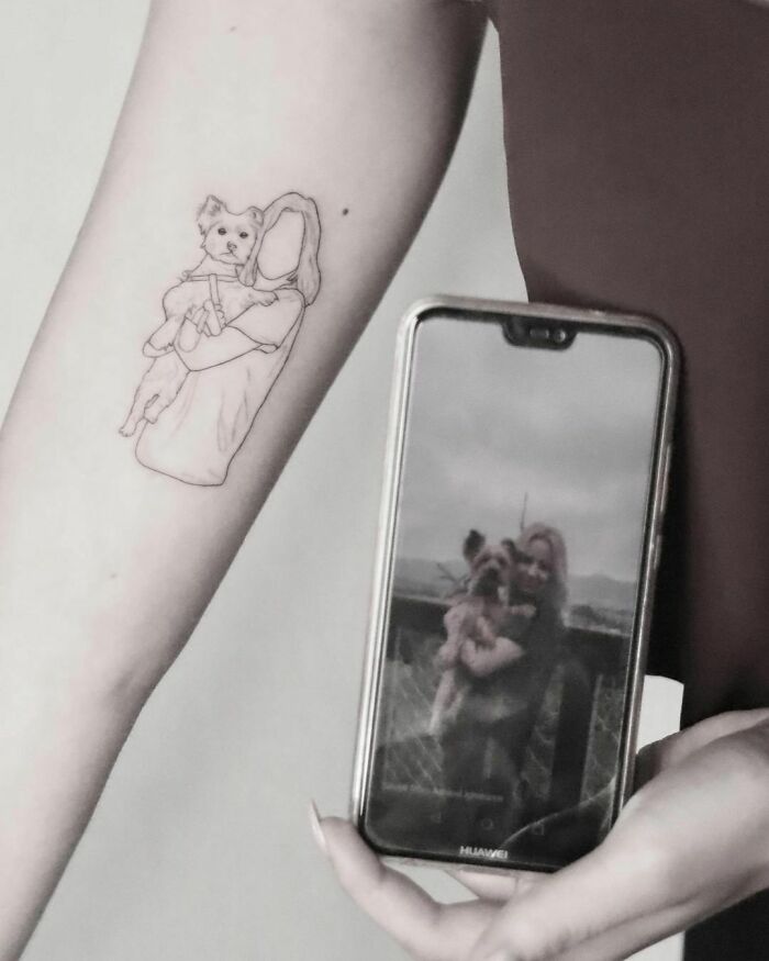 Pet tattoo of a person holding a dog on the forearm, with a matching photo displayed on a smartphone.