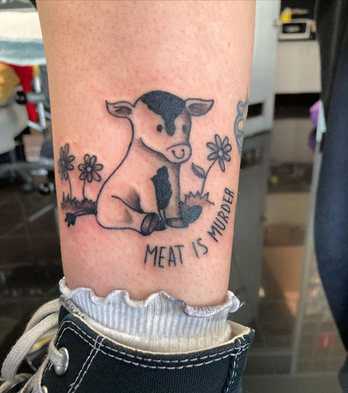 Animal tattoo of a cute cow with flowers and the phrase "Meat is M****r" on a person's leg.