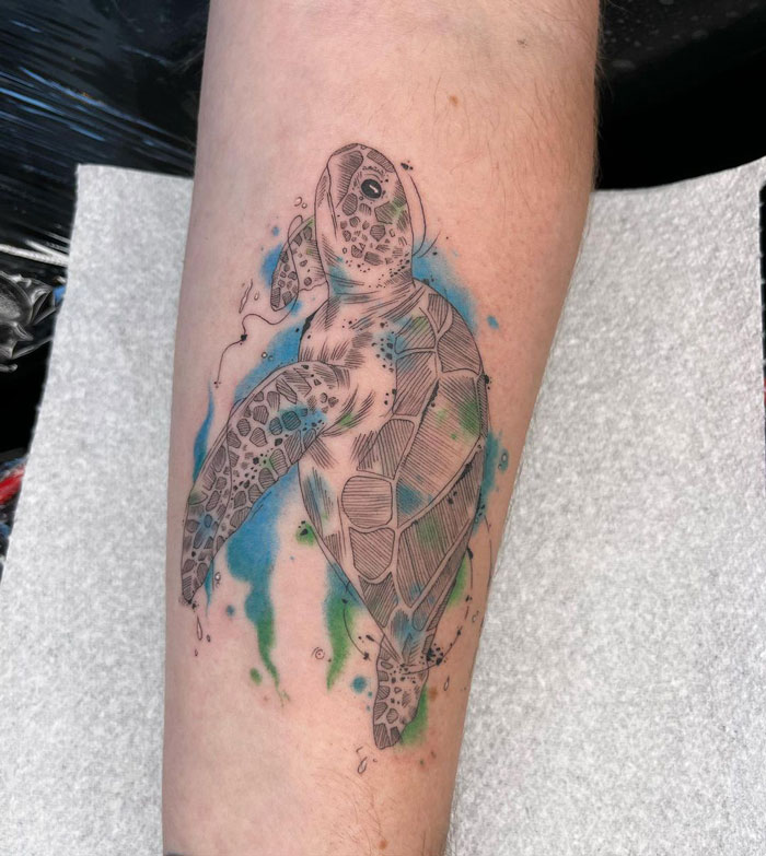Animal tattoo of a sea turtle on forearm with watercolor splash background.