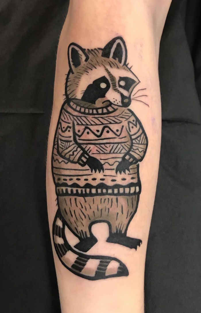 Animal tattoo of a raccoon in a patterned sweater, blending simplicity and realism.