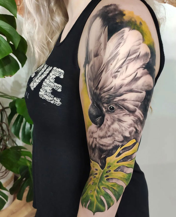 Parrot tattoo on woman's arm, featuring detailed feathers and green leaves, celebrating the animal kingdom.