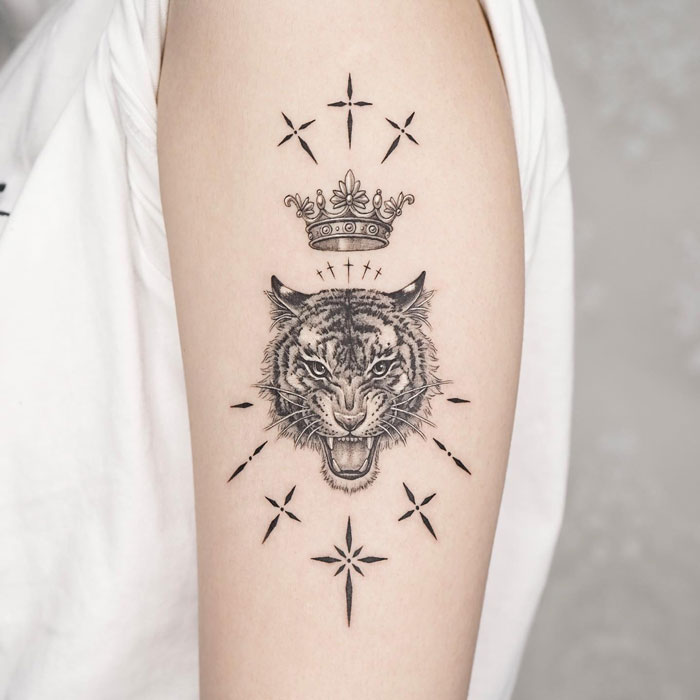Animal tattoo of a roaring tiger with a crown on an arm, surrounded by star-like patterns.