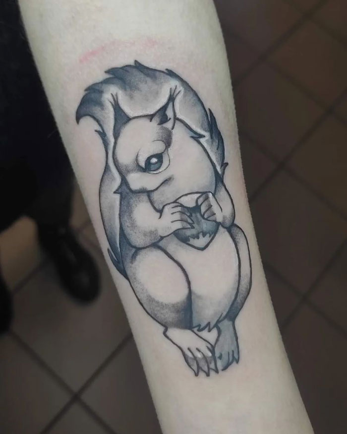 Animal tattoo of a cartoon-style squirrel holding an acorn on a forearm.
