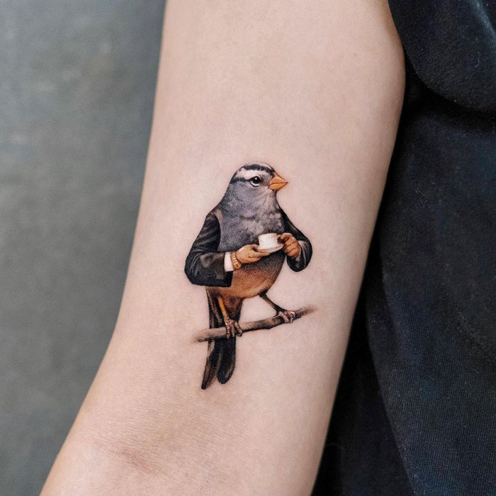 Animal tattoo of a bird in a suit holding a cup on a person's arm, showcasing creative design ideas.