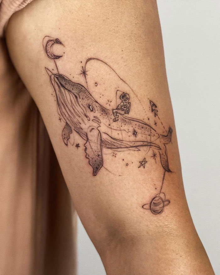 Minimalist animal tattoo of a whale with an astronaut, surrounded by space elements on a person's arm.