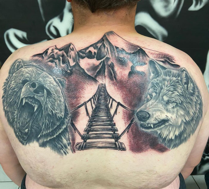 Back tattoo featuring a roaring bear, a wolf, a mountain landscape, and a suspension bridge; animal tattoo inspiration.