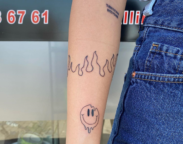 Arm with a flame armband tattoo design above a melting smiley face tattoo.