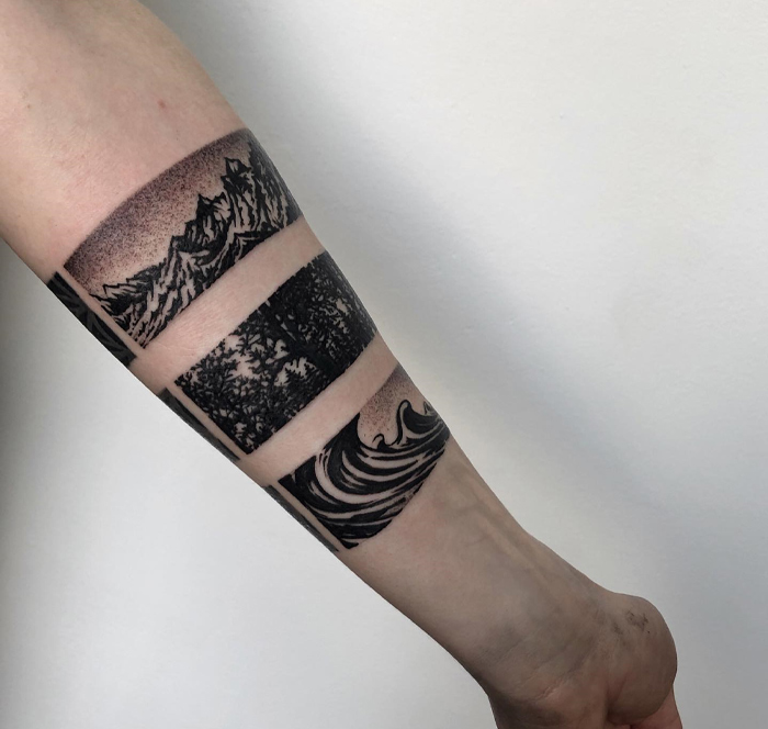 Armband tattoo featuring mountain, forest, and wave designs in black ink on a forearm.
