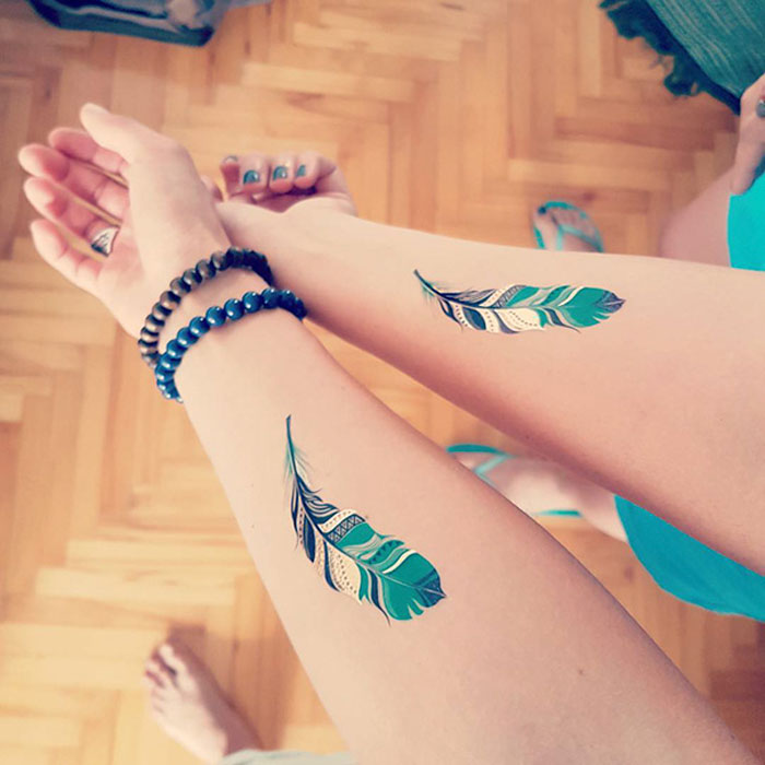 Two forearms with matching feather tattoos symbolize best friend tattoos, adorned with beaded bracelets.