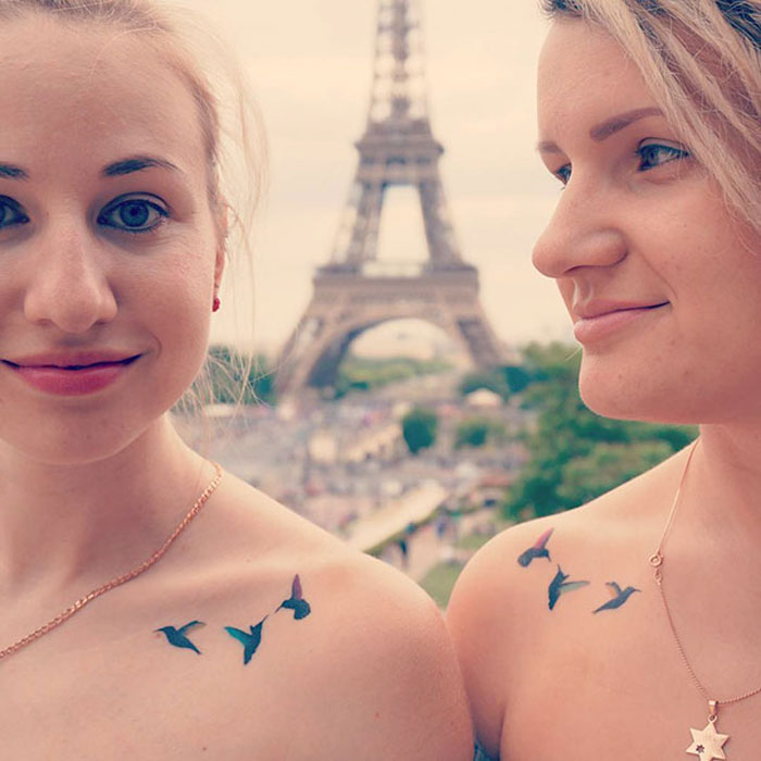 Two friends with matching bird tattoos on their shoulders, standing near the Eiffel Tower, celebrating their friendship.