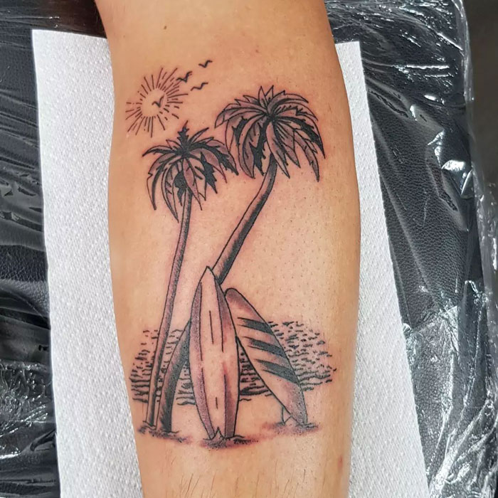 Lower leg tattoo featuring palm trees and surfboards on a beach under the sun.