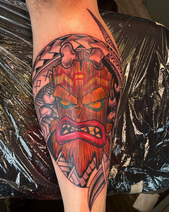 Colorful tiki mask calf tattoo with bold geometric patterns.
