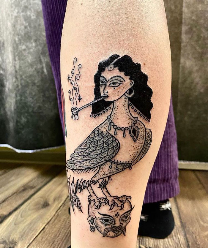 Lower leg tattoo of a mythical creature with a woman's face, detailed patterns, and artistic elements.