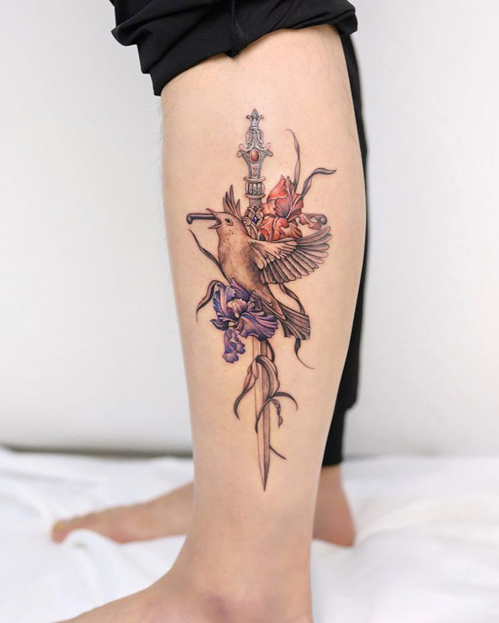 Calf tattoo with a bird and flowers around a dagger, showcasing intricate design and vibrant colors.