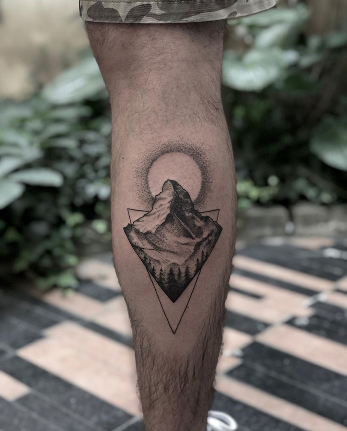 Mountain design calf tattoo with geometric shapes and a sun halo in a natural setting.