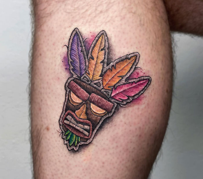 Colorful tribal mask tattoo on lower leg with vibrant feathers.