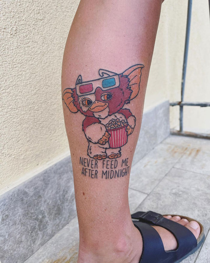 Calf tattoo featuring a cute creature with 3D glasses holding popcorn, under the phrase "Never feed me after midnight".