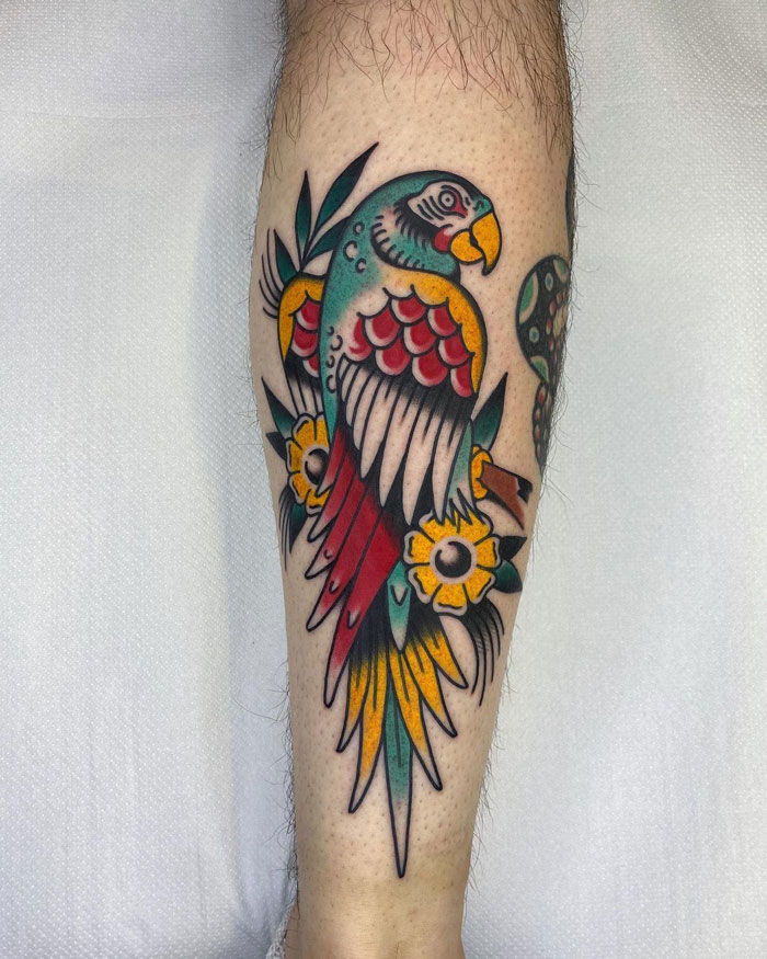 Colorful parrot tattoo on lower leg, featuring vibrant reds, greens, and yellows.