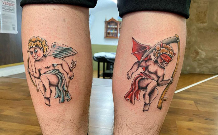 Lower leg tattoos of angel and devil cherubs with wings and spears on a wooden-floor background.