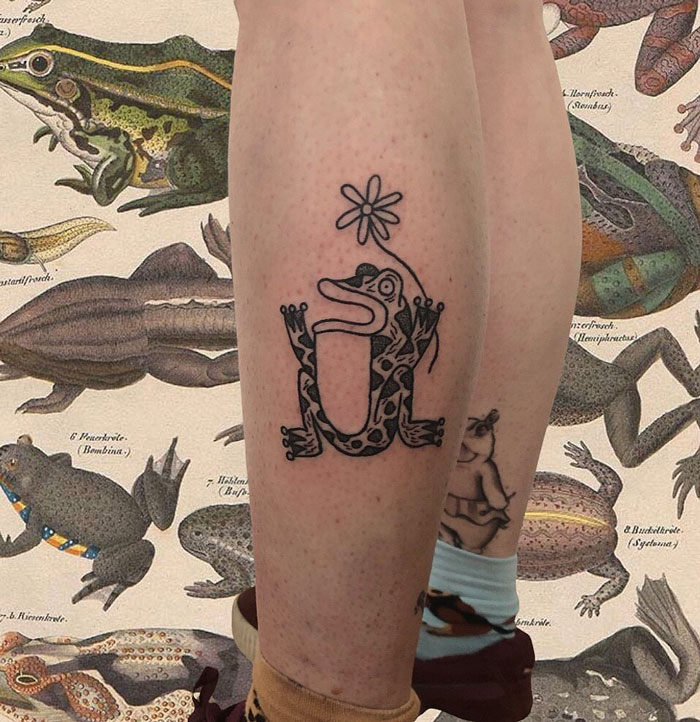 Lower leg tattoo of a whimsical frog design with a flower on a person's calf, set against an illustrated frog background.