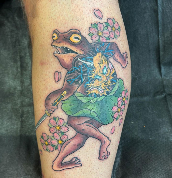 Tattoo of a frog with a sword on a calf, surrounded by cherry blossoms, showcasing cool tattoo art.