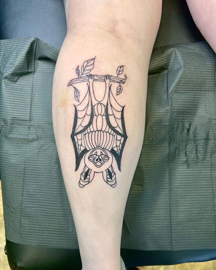Lower leg tattoo of a stylized bat hanging upside down with leaves on a branch.