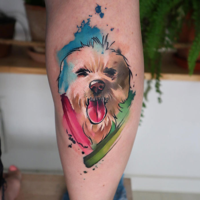 Watercolor-style lower leg tattoo of a dog's face with multicolored splashes.