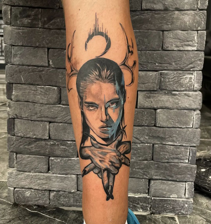 Unique calf tattoo featuring a stylized face with antlers and intricate detailing, set against a brick background.