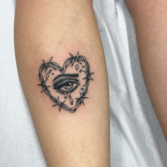 Heart-shaped eye tattoo with barbed wire on lower leg.