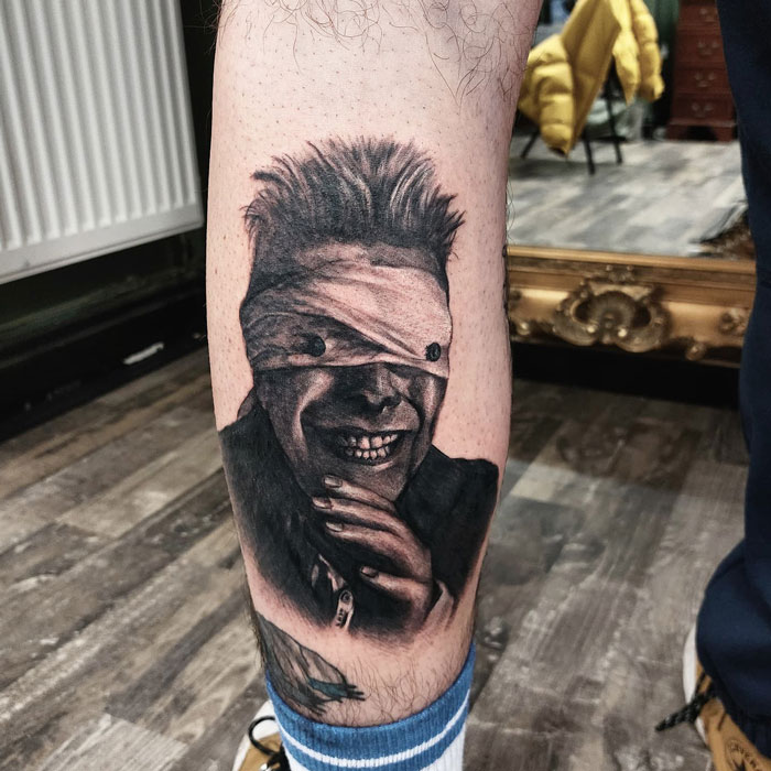 Unique calf tattoo of a grinning figure with eyes covered, showcasing intricate detailing and shading.