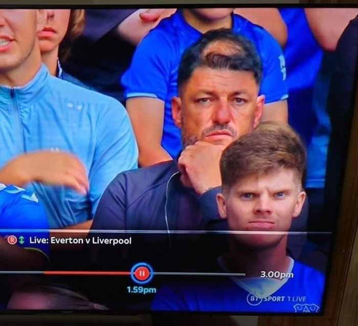 Fans at a soccer match, with one sporting an unusual bad haircut, Everton vs Liverpool game on TV screen.