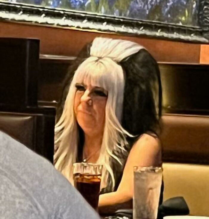 Person with a striking bad haircut, featuring a voluminous black and white style, sitting at a booth in a restaurant.